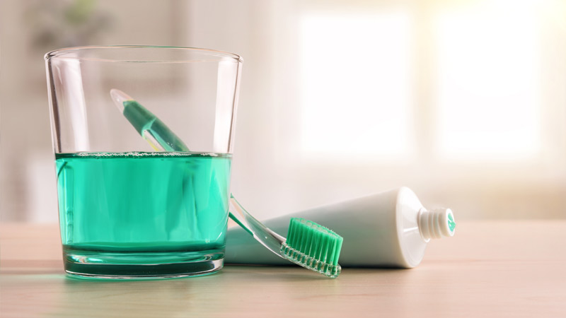 Copo com elixir de cor verde ao lado de pasta e escova de dentes, em cima de uma mesa